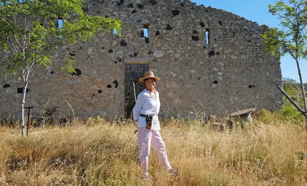 Pascale Rivière, vigneronne, portant un chapeau de paille, debout devant une ancienne ruine en pierre dans un champ, avec des arbres et des herbes sèches autour