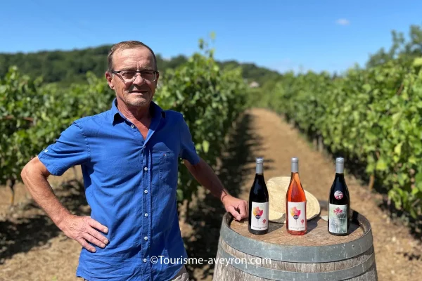 Jean-Marie Paillard portant une chemise bleue et des lunettes, debout à côté d'un tonneau en bois avec trois bouteilles de vin et un chapeau de paille, dans les rangées de vignes verdoyantes sous un ciel bleu clair