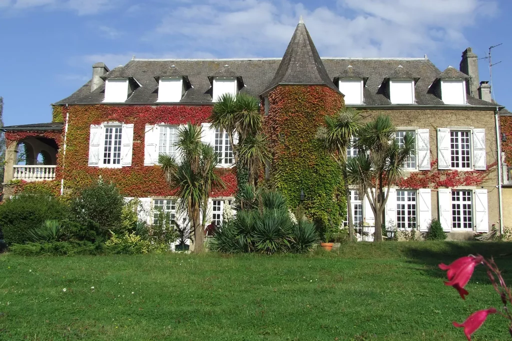 Grande maison du domaine de Cuqueron avec une façade recouverte de vigne vierge rouge et verte, entourée de palmiers et de jardins verdoyants, sous un ciel bleu partiellement nuageux