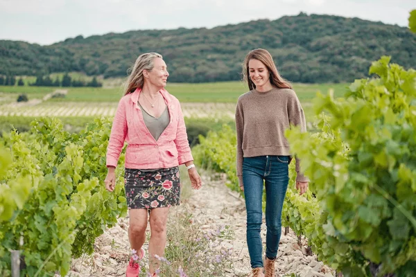 Madeleine et Ambre marchent côte à côte dans les rangées de vignes, discutant et souriant, avec un paysage de collines verdoyantes en arrière-plan
