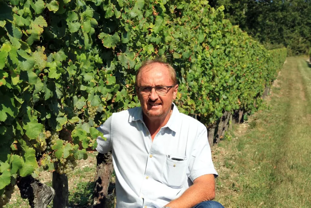 Vincent Vesselle, vigneron du Château La Rayre, accroupi à côté de rangées de vignes verdoyantes, portant une chemise blanche et des lunettes, par une journée ensoleillée