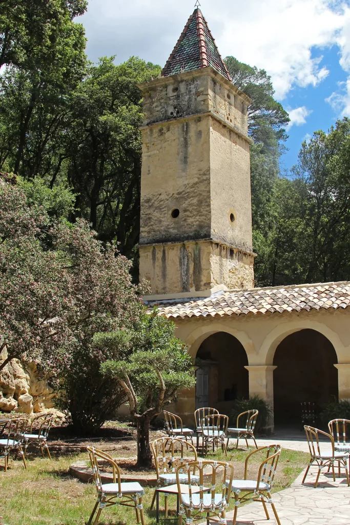 Vue extérieure d'un bâtiment ancien du Château de l'Olivète avec une tour en pierre et un toit en tuiles, entouré de chaises disposées sur une pelouse. Le bâtiment est partiellement ombragé par des arbres