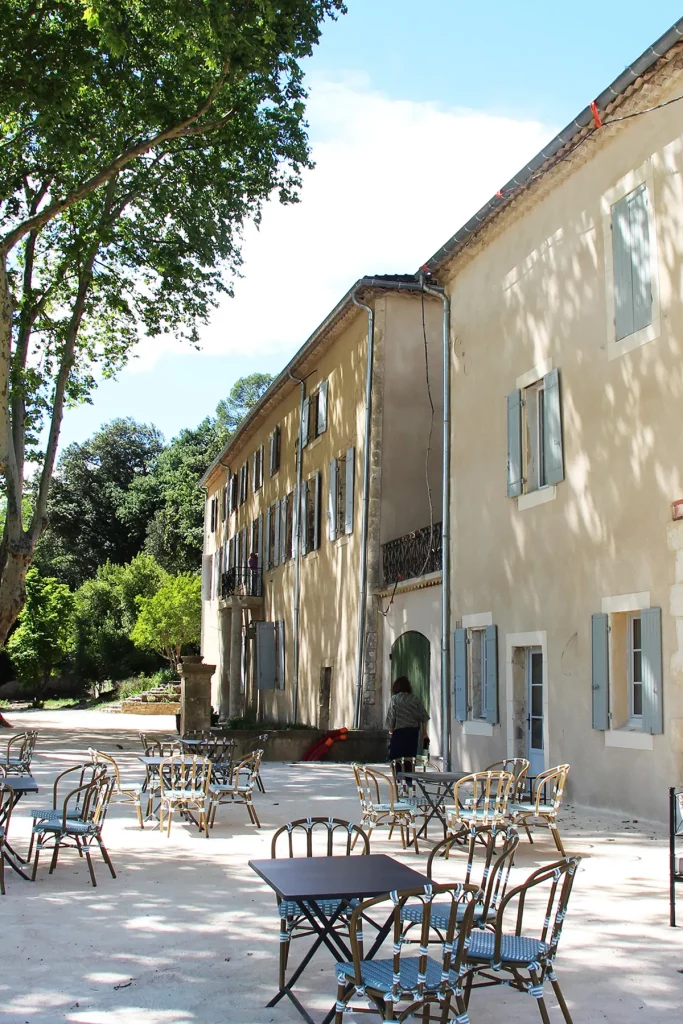 Vue extérieure d'une bâtisse du Château de l'Olivète, avec ses volets bleus, entourée de tables et de chaises en plein air. Les arbres projettent de l'ombre sur la cour