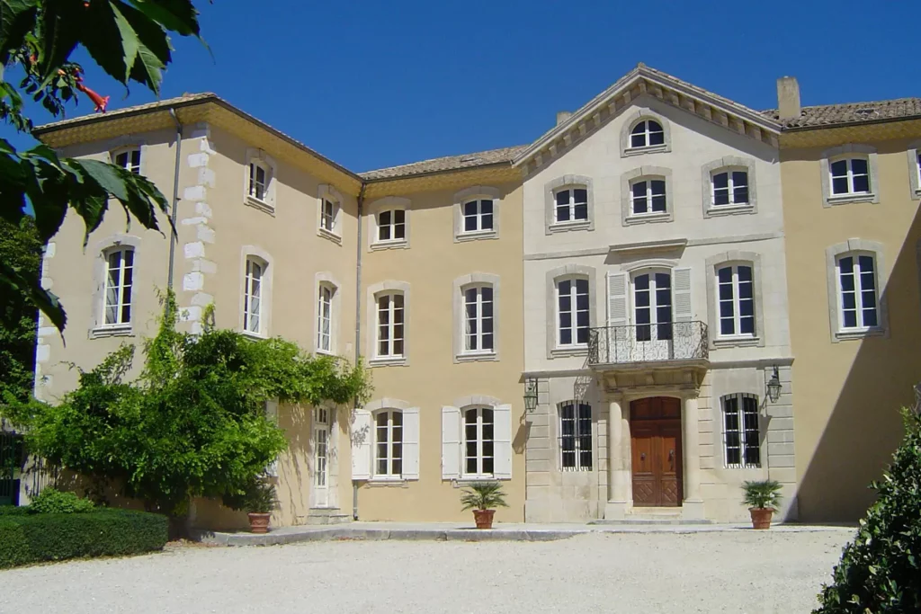 Façade principale du château Rochecolombe avec ses murs de couleur beige, ses nombreuses fenêtres blanches et son balcon central. La cour est ornée de plantes vertes et le ciel est bleu clair en arrière-plan.