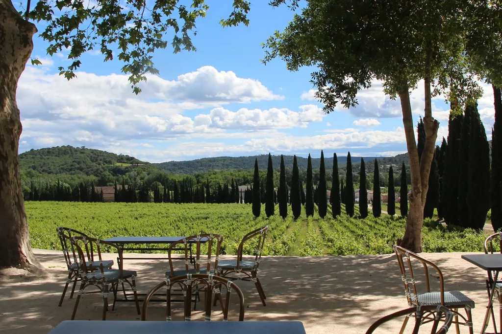 Vue depuis la terrasse du Château de l'Olivète avec des tables et des chaises à l'ombre des arbres, surplombant les vignes et les cyprès en arrière-plan, sous un ciel bleu avec des nuages blancs