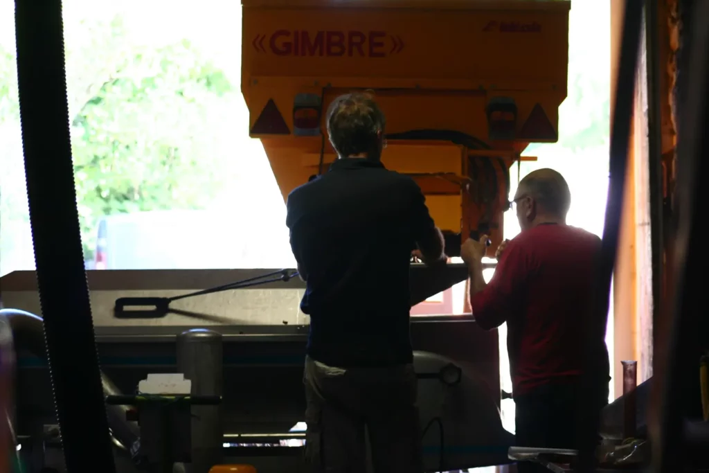 Deux vignerons de dos travaillant sur une machine de récolte de raisins, éclairés par une lumière naturelle provenant de l'extérieur